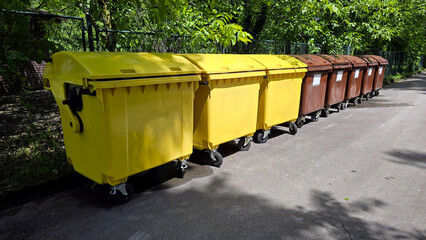 A row of large yellow and brown waste bins with lids and wheels stands along a sunlit street, bordered by a metal fence and leafy green trees, illustrating organized urban waste management