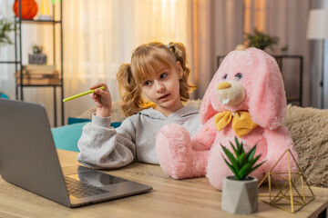 Young school girl sitting with plush bunny toy near laptop, watching an educational video online at home. Caucasian child pauses the video to explain material to rabbit friend with joy and curiosity
