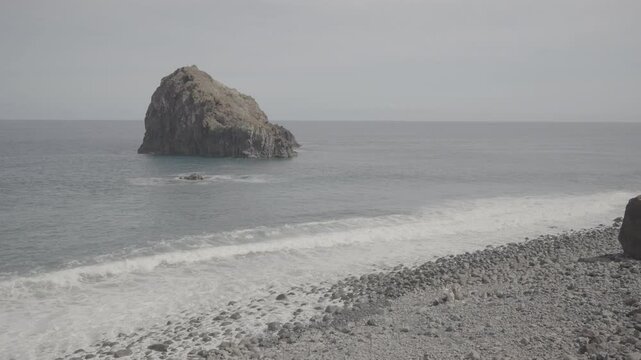 Scenic pebble bay facing rugged islet amid gentle surf off madeira (SLOG3)