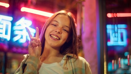 Joyful Caucasian Asian girl showing two fingers gesture in city junior happy teen smile teenager...