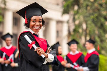 Cheerful african american young lady in graduation costume showing her diploma and smiling at...