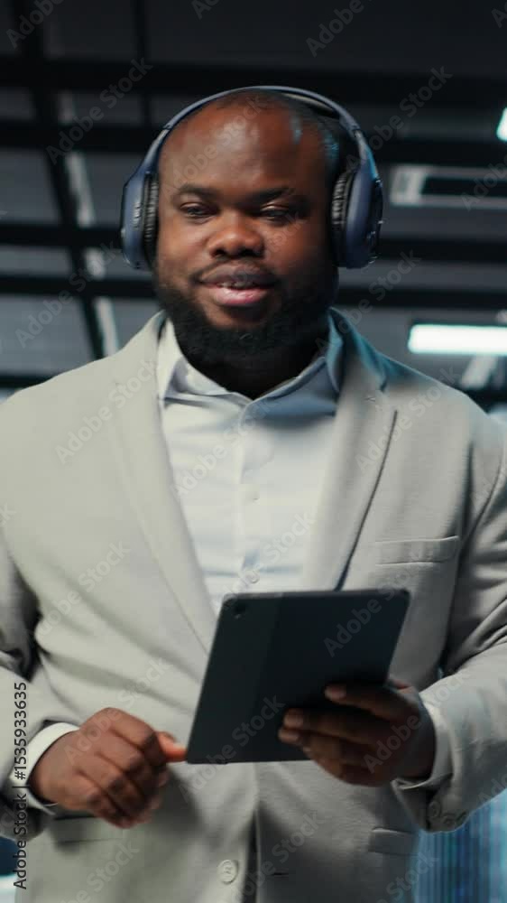 Poster vertical video data center admin wearing headphones using tablet to identify bottlenecks. server far