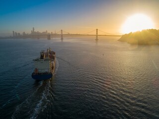 Container ship sails San Francisco Bay past the city skyline and Bay Bridge at sunset in...