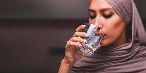 Portrait Of Arab Woman Drinking Water From Glass Standing In Kitchen Indoors, Wearing Hijab...