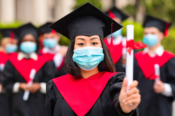 Asian young lady student in graduation costume and protective face mask posing over international...