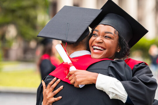 Congratulations. Happy african american couple students in graduation dresses and hats embracing, having ceremony at university campus, enjoying and celebrating graduation, closeup portrait