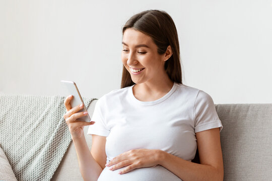 Happy pregnant millennial woman using smartphone at home, browsing pregnancy app while sitting on couch in living room, smiling expectant lady looking at phone screen and touching belly, free space - Powered by Adobe