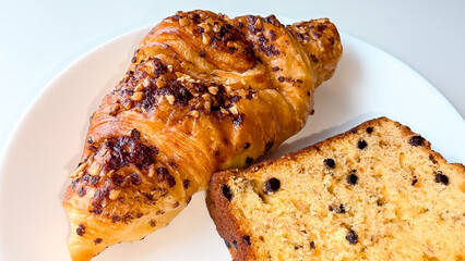 Delicious close-up of a croissant with chopped nuts and a slice of chocolate chip banana bread on a white plate. Perfect for bakery promotions, breakfast menu designs, or food blog visuals.