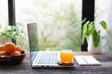 Side view of a laptop with fresh orange juice on rustic wooden table, by the window