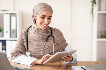 Smiling attractive female doctor woman in hijab using digital tablet in office, sitting at workdesk...