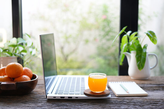 Side view of a laptop with fresh orange juice on rustic wooden table, by the window