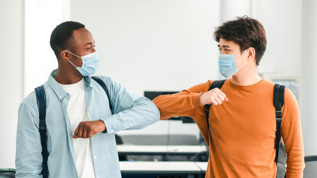 Elbow Bump. Smiling multicultural students wearing medical face masks greeting each other at classroom. Asian and african american men avoid touching, studying in university, standing at lecture hall