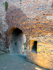 Fototapeta premium Narrow medieval alley framed by ancient brick walls, featuring an arched tunnel and small window niche. Soft lighting enhances the aged texture of this fortress corridor.