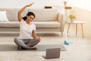 Training At Home. Active young black woman practicing yoga in side bend pose, stretching arm and watching tutorial training on laptop in the morning, sitting on mat in bedroom, free space