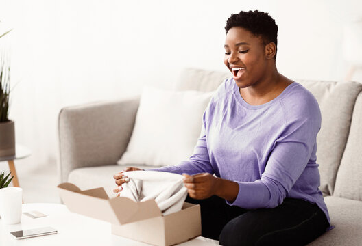 Happy Customer Concept. Portrait of excited curvy black lady opening and unpacking cardboard box parcel, sitting on the couch in living room at home. Young happy female satisfied with delivery service