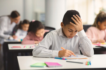 Education And Learning Concept. Portrait of focused small boy sitting at desk in classroom at school, writing in notebook, having test or exam and thinking, touching and grabbing head