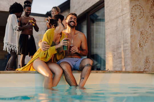 Young couple kissing and enjoying drinks at luxury pool party with friends