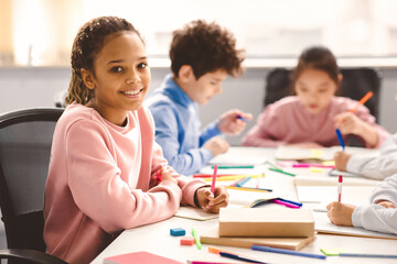Academic Concept. Smiling junior african american school girl sitting at desk in classroom, writing in notebook, posing and looking at camera. Group of classmates studying in the background