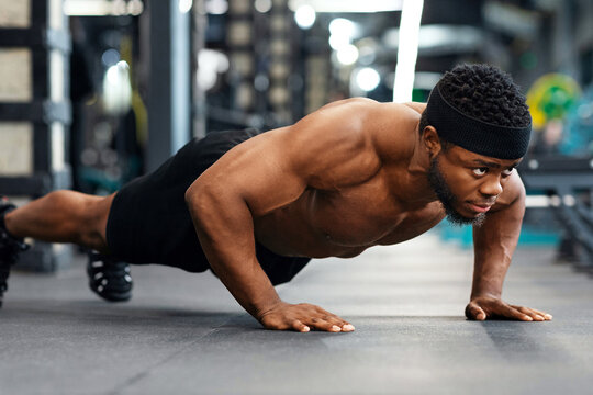 Push-ups, press-ups workout. Muscular african american shirtless guy sportsman building up muscles, young black man training on floor at modern gym, doing push-ups, panorama - Powered by Adobe