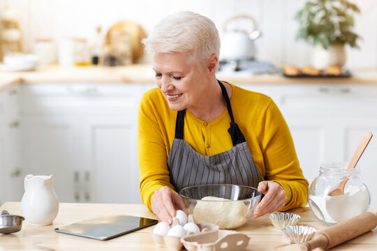 Smiling aged lady cooking in kitchen, looking at digital tablet, watching online culinary class while kneading dough, copy space. Happy old woman cooking by recipes from Internet, using pad