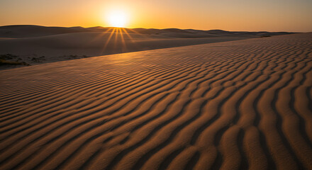 Rippled sand dunes in the desert with a beautiful sunburst at sunrise or sunset on the horizon. A vast, warm, and serene natural landscape.
