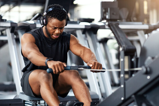 Motivated young black sportsman with wireless headset having workout on rowing machine. Handsome african american man bodybuilder training body on modern block exerciser in gym, copy space - Powered by Adobe
