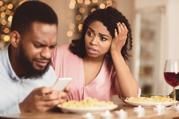 Phubbing. Angry young black woman looking at her boyfriend stuck in smartphone, unhappy about neglect and lack of communication. Jealous lady and gadget addicted man having dinner in cafe or at home