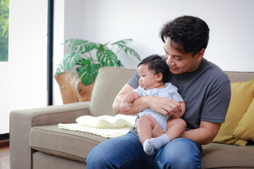 Asian father holding a little baby in his arms. He smiles happily sitting on the sofa at home. The bond between father and baby helps the baby's development. Family concept. Father's day.