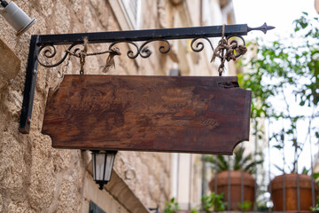 A rustic wooden sign hangs empty from an ornate iron bracket in a charming vintage street. the backdrop features stone walls and potted plants, creating a quaint atmosphere.