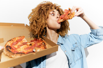 Young woman with curly hair enjoying pizza from a takeaway box, expressing joy and indulgence, ideal for food lovers and healthy eating concepts