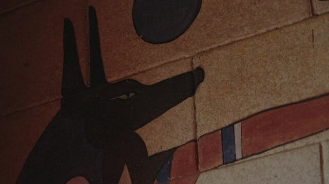 Ancient Egyptian mural painting of Anubis, the jackal-headed god of the dead. The artwork depicts Anubis against a textured, light brown wall, possibly within a tomb, ancient Egyptian