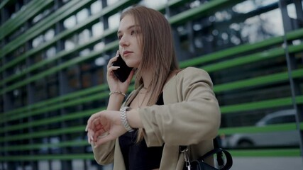 Portrait of a focused young woman checking her watch while making a phone call, standing in an urban environment with modern architecture. Young Woman Checking Time While Talking on Phone in City - Powered by Adobe