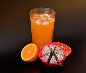 Fruit juice in a tall glass with ice on a black background, halves of ripe mango, orange and grapefruit nearby.