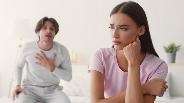 In a contemporary bedroom, a man expresses frustration while a woman looks upset and distant. The tension indicates unresolved issues between the couple in their personal space.
