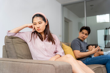 Fototapeta premium New Mother Feeling Stressed or Unhappy, Partner Distracted, Couple with Newborn Experiencing Marital or Postpartum Issues, Unhappy Woman Sitting Apart from Partner and Baby on Sofa