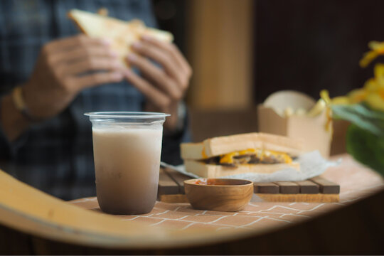 A plastic cup of iced coffee served on a table with sandwiches, chocolate bars, and a small wooden bowl of sauce. In the background, a person wearing a plaid shirt is holding and eating a sandwich,