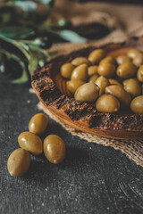 Rustic wooden bowl filled with green olives on burlap fabric with olive branch in background. Natural warm lighting and dark wood backdrop add to the organic Mediterranean atmosphere