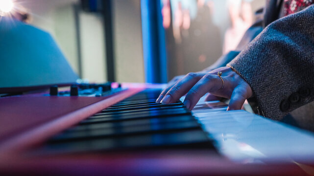 Close-up of hands playing electronic keyboard during live music performance with blurred bassist in background under blue lighting