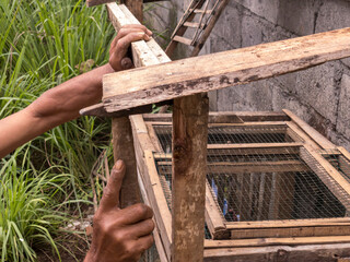 A man repairs a bamboo chicken cage with a hammer. The rustic setting highlights traditional craftsmanship and rural life. © Ndorography