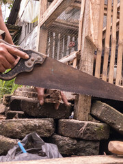 A man using a rusty handsaw to repair a bamboo cage on a stone floor. This scene captures traditional manual labor and rural craftsmanship in Indonesia.