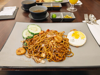 Close-up of a delicious plate of Malaysian-style fried noodles (Mee Goreng Mamak) served with fried egg, prawns, squid, cucumber slices, and sambal. Local cuisine on a modern table setting