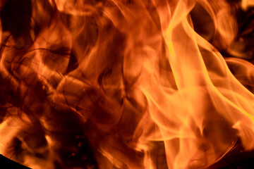 Close-up image of bright orange flames against a backdrop of black ash and burnt debris. A strong...