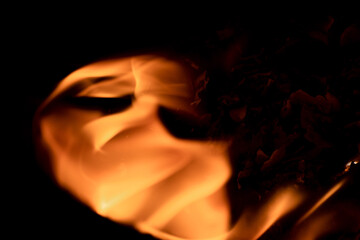Close-up image of bright orange flames against a backdrop of black ash and burnt debris. A strong...