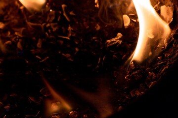 Close-up image of bright orange flames against a backdrop of black ash and burnt debris. A strong...