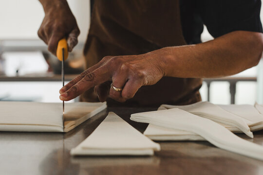 Close-up of a baker’s hands carefully rolling croissant dough on a metal countertop. The person is wearing a brown apron and has flour on their fingers, creating an authentic, artisanal atmosphere in 