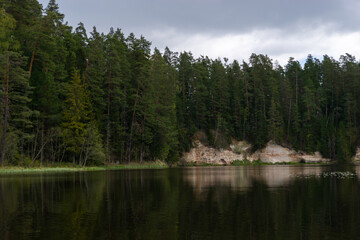 Fototapeta premium Mossy Cliffside with Dark Cave Entrance and Reflections in Calm Forest River Surrounded by Evergreen Trees