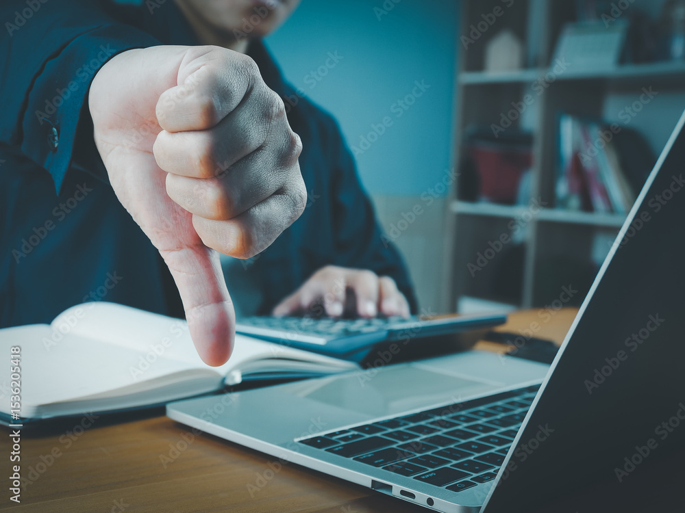 Poster person showing thumbs down gesture in front of laptop and notebook, expressing disapproval or dissat