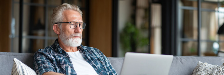 Older man with glasses and beard sitting on couch using laptop indoors