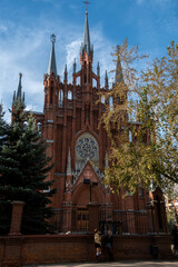 Stunning view of a historic red brick cathedral with elegant spires rising against a clear blue sky. Seen from a tree-lined street with lush greenery enhancing the serene atmosphere.