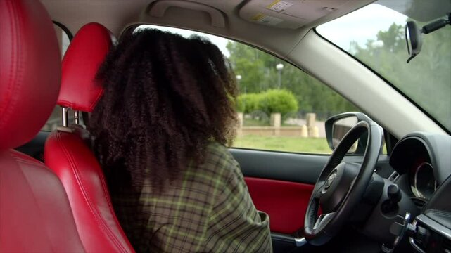 Beautiful African American female driver in casual clothes entering car, buckling up seat belt and pushing engine start button while driving vehicle in city during road trip.